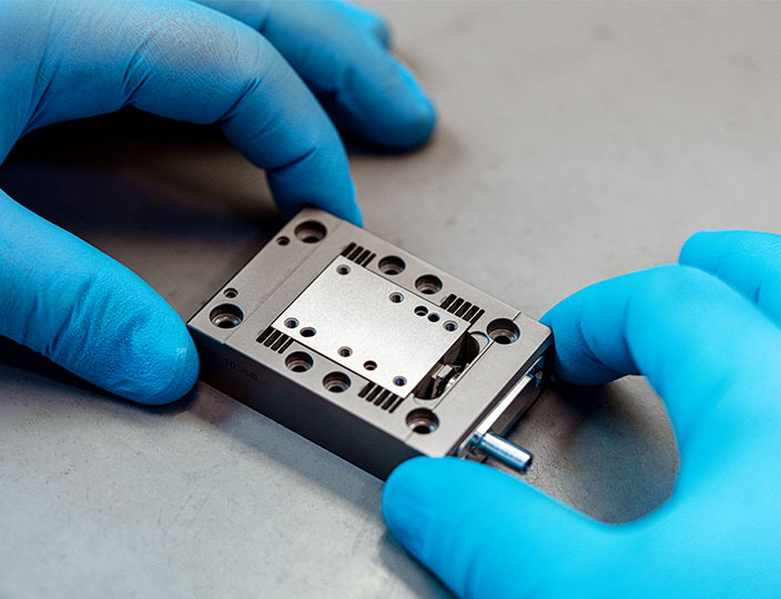 An operator's blue-gloved hands hold a Queensgate piezo nanopositioning stage on a grey mat.